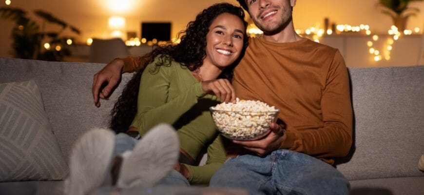 A man and a woman sitting on a living room couch with a bowl of popcorn