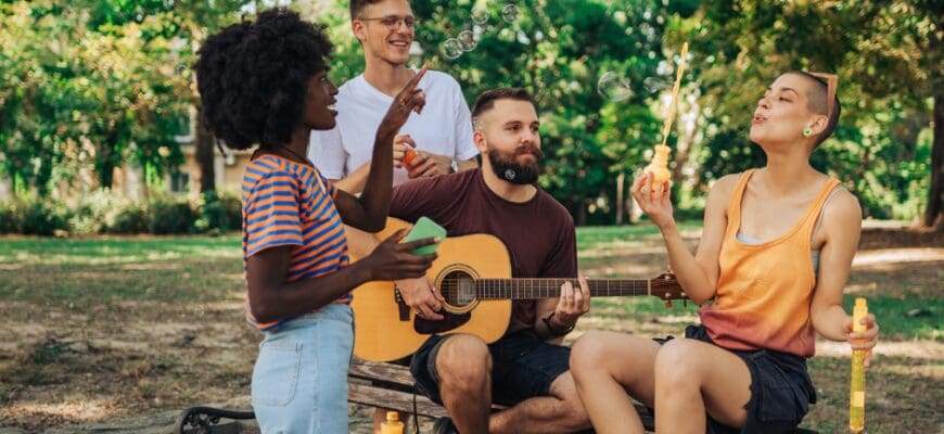 Four people gathered around an outdoor park bench, one holding a guitar